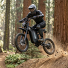 A rider wearing full protective gear jumps the HappyRun pedal assist Electric Dirt Bike G18 Pro over a dirt trail in a forest
