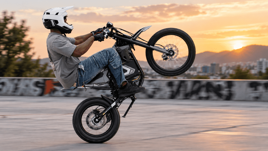 Electric dirt bike with a rider performing a wheelie against a sunset cityscape.