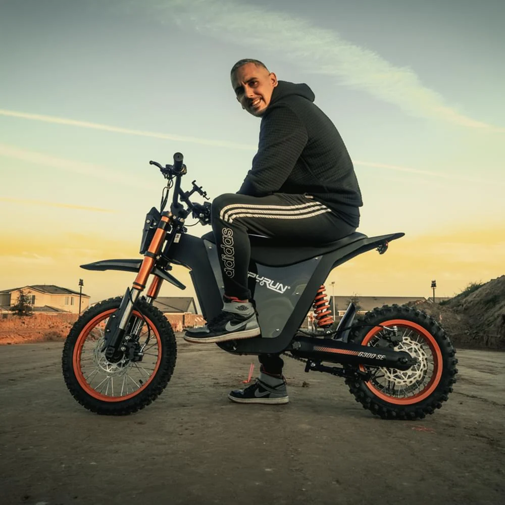HappyRun G300 SE electric dirt bike with full suspension and knobby off-road tires, rider seated on the bike outdoors at sunset with a residential backdrop