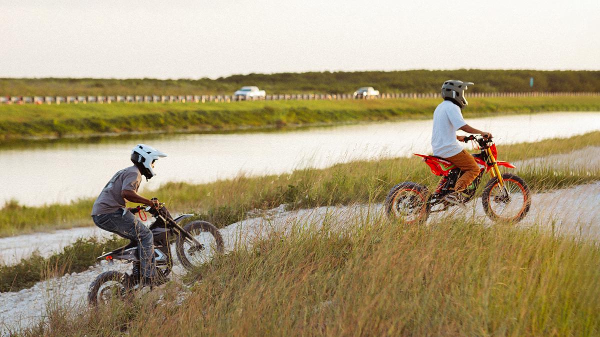 Two riders on electric pit bike and dirt bike riding along a sandy waterside trail
