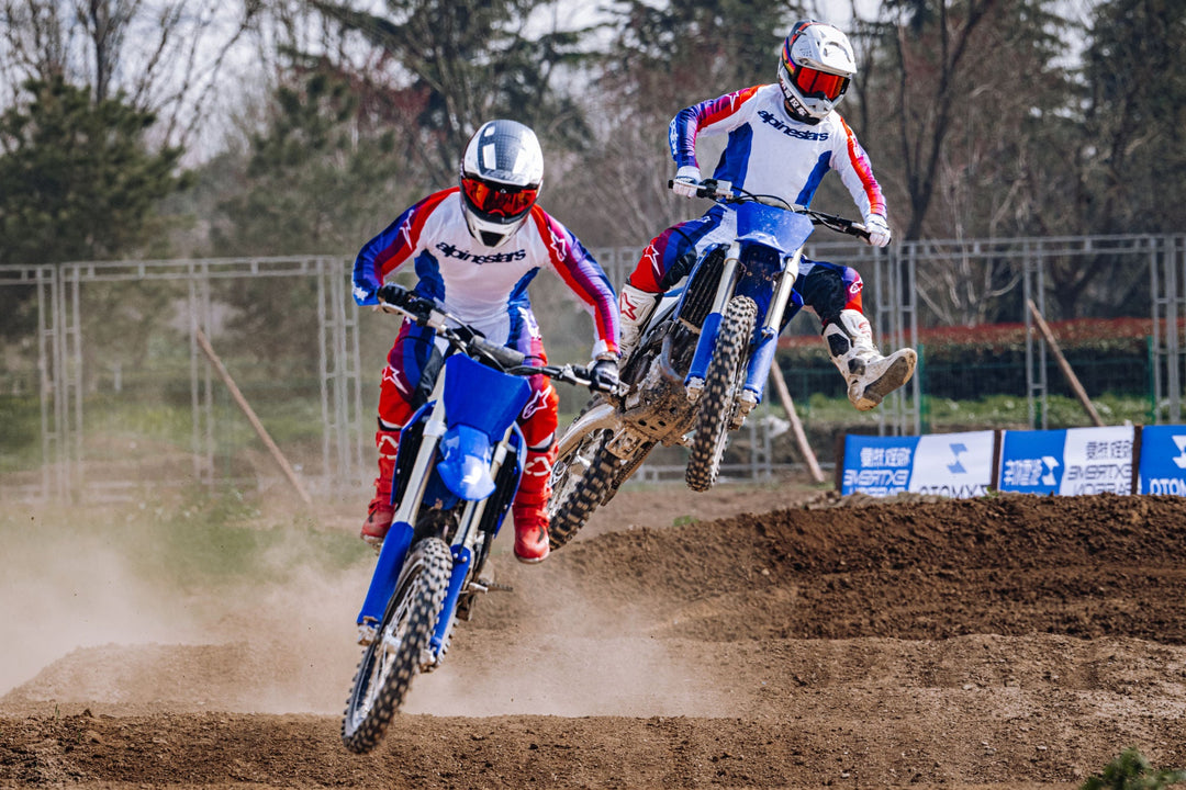 Two riders on blue ZXMOTO dirt bikes, with one airborne over a dirt jump behind the other